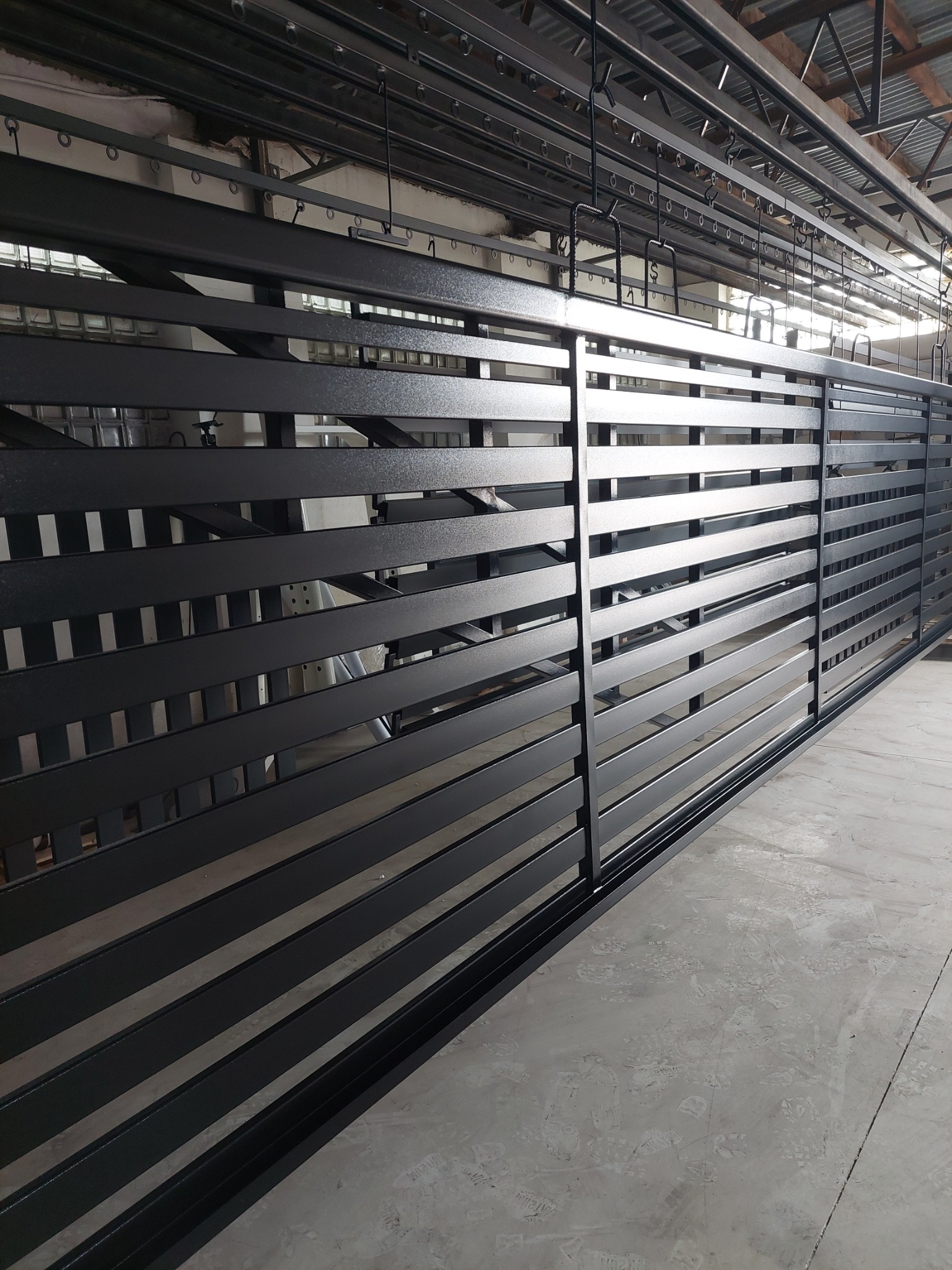 Metalowa brama przesuwna z poziomymi szczeblami, świeżo pomalowana na czarno, zawieszona na hakach w hali produkcyjnej, widoczne ślady butów na betonowej podłodze.