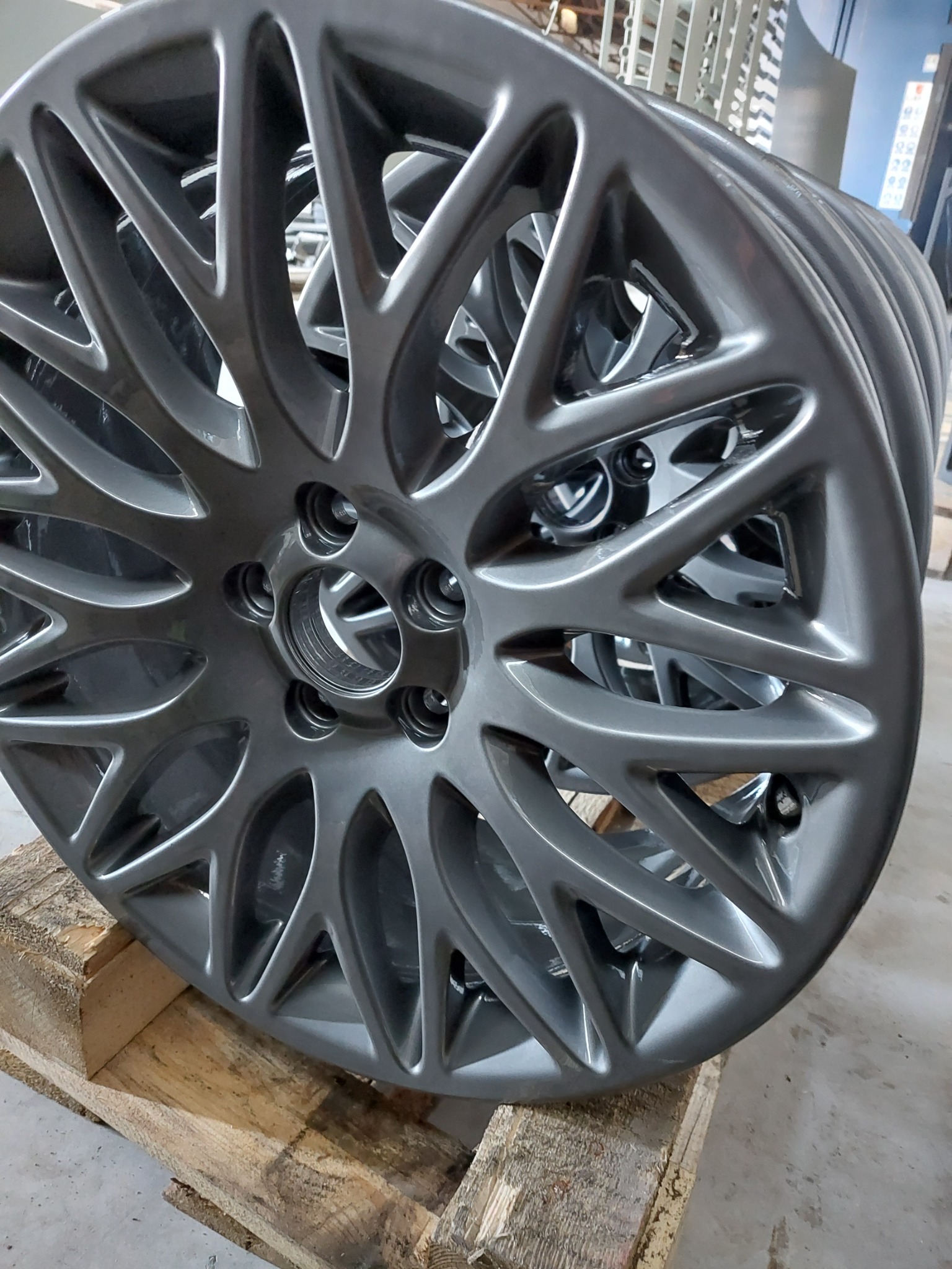 Stos szarych felg aluminiowych, świeżo po nałożeniu powłoki, ułożony na drewnianej palecie w hali produkcyjnej.