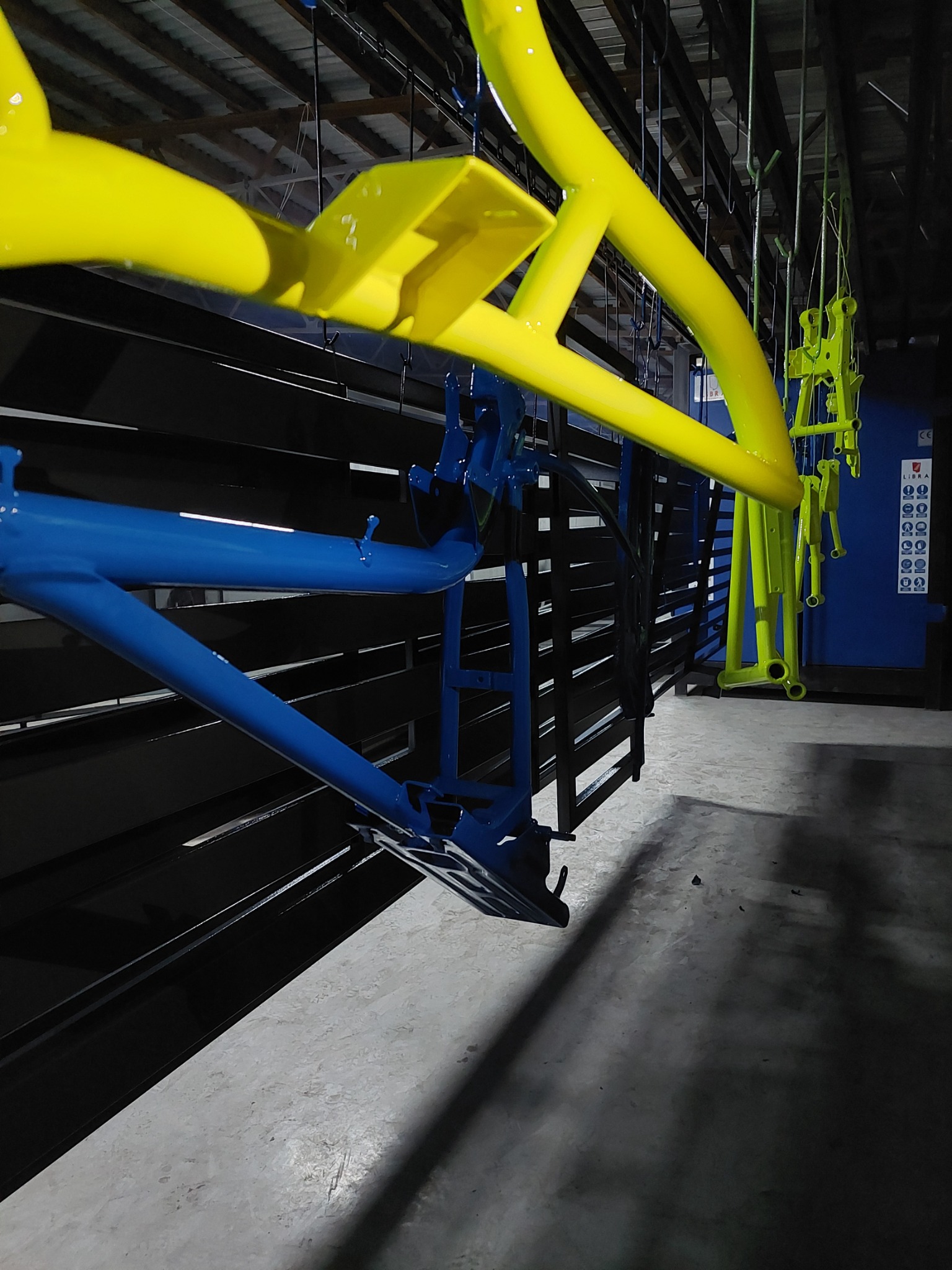 Ramy rowerowe w kolorze niebieskim i żółtym po malowaniu proszkowym, zawieszone na wieszakach w hali produkcyjnej przed piecem.