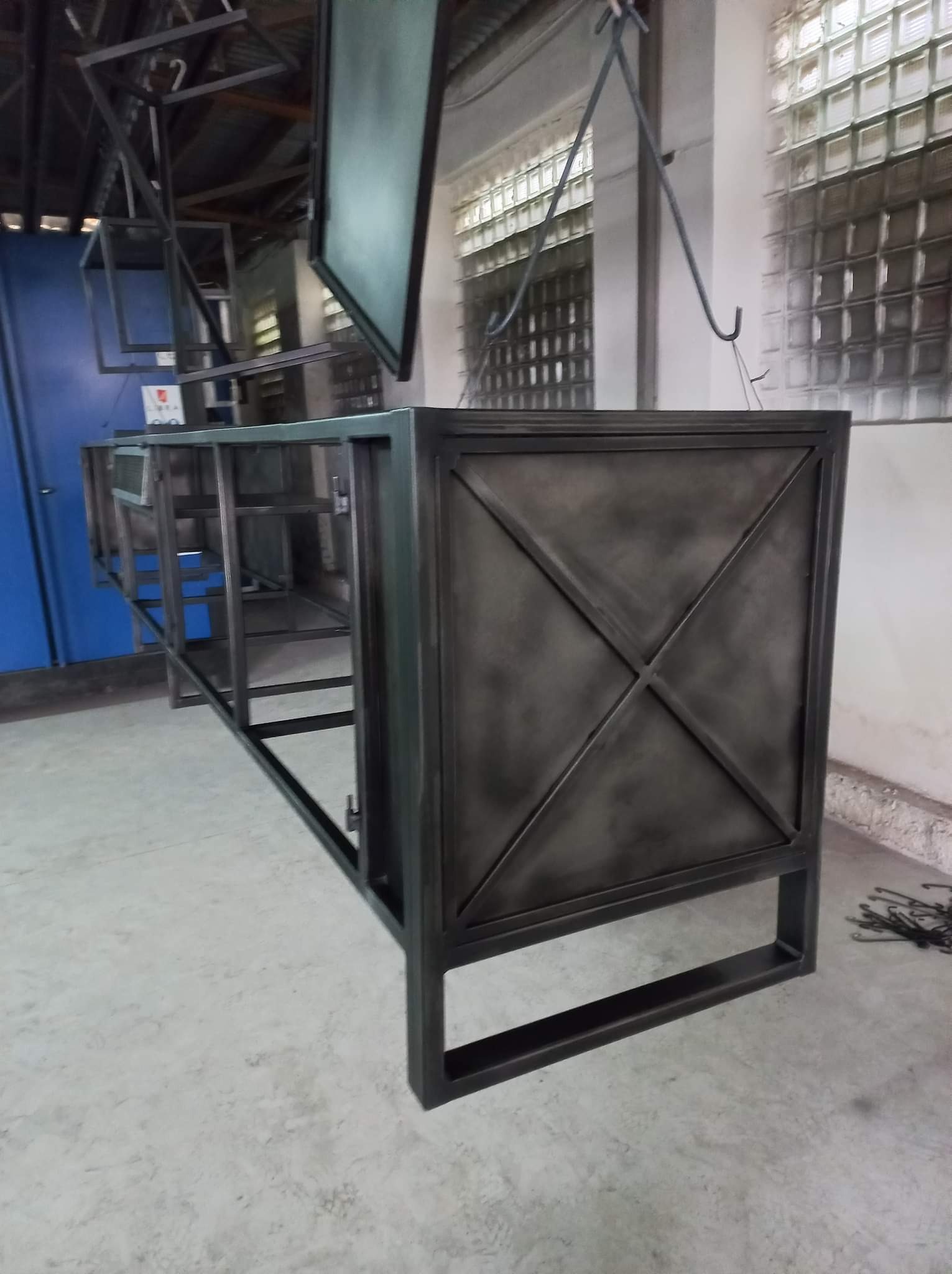 Metalowa konstrukcja meblowa w trakcie obróbki, zawieszona na hakach w warsztacie, widoczne ślady po obróbce powierzchniowej i surowy wygląd metalu.