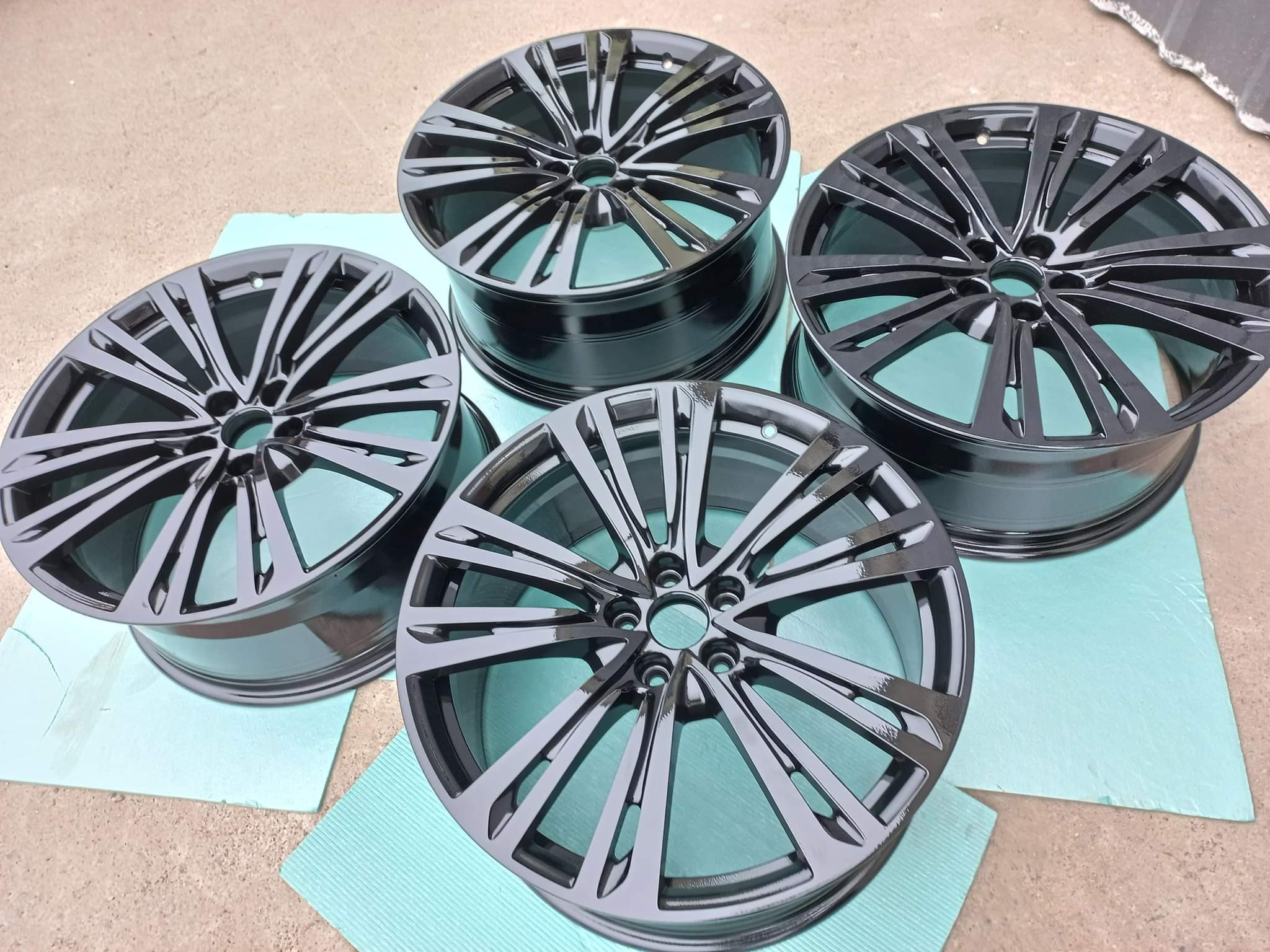 Cztery pomalowane na czarno felgi samochodowe, ułożone na niebieskiej macie, prezentują połyskującą powierzchnię i nowoczesny design.