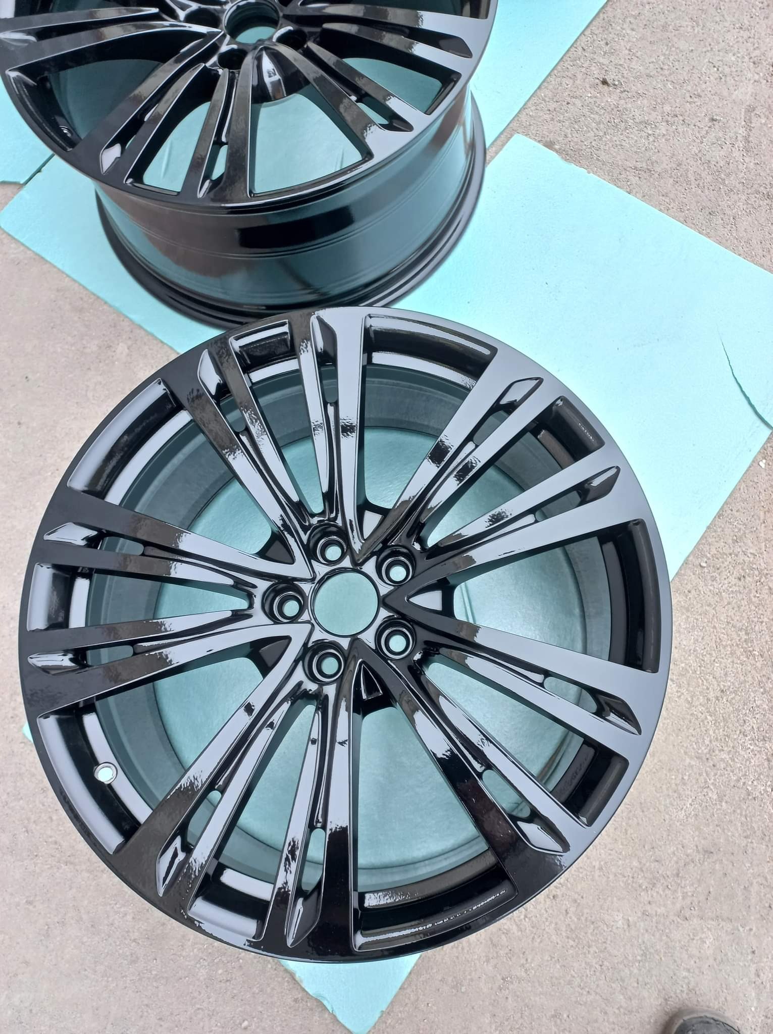 Dwa aluminiowe felgi samochodowe po renowacji, pomalowane na wysoki połysk w kolorze czarnym, leżą na niebieskiej folii ochronnej na szarym betonowym podłożu.