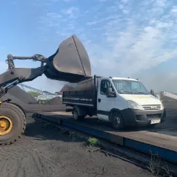 Ładowarka czołowa z łyżką nasypuje węgiel do skrzyni białego samochodu dostawczego z plandeką, widoczny napis 'Transport - Wywrotka 3,5t'.