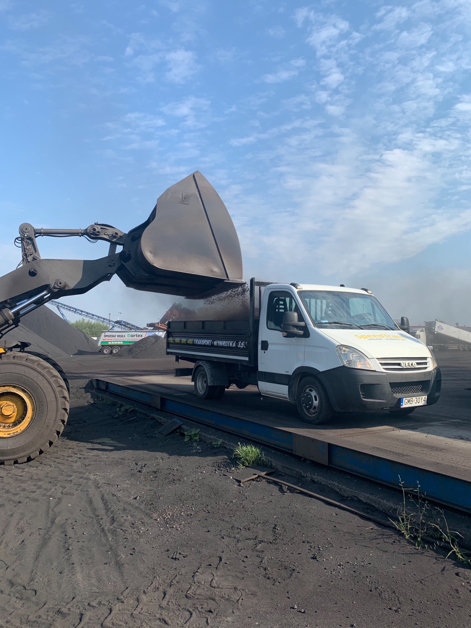 Ładowarka czołowa z łyżką nasypuje węgiel do skrzyni białego samochodu dostawczego z plandeką, widoczny napis 'Transport - Wywrotka 3,5t'.