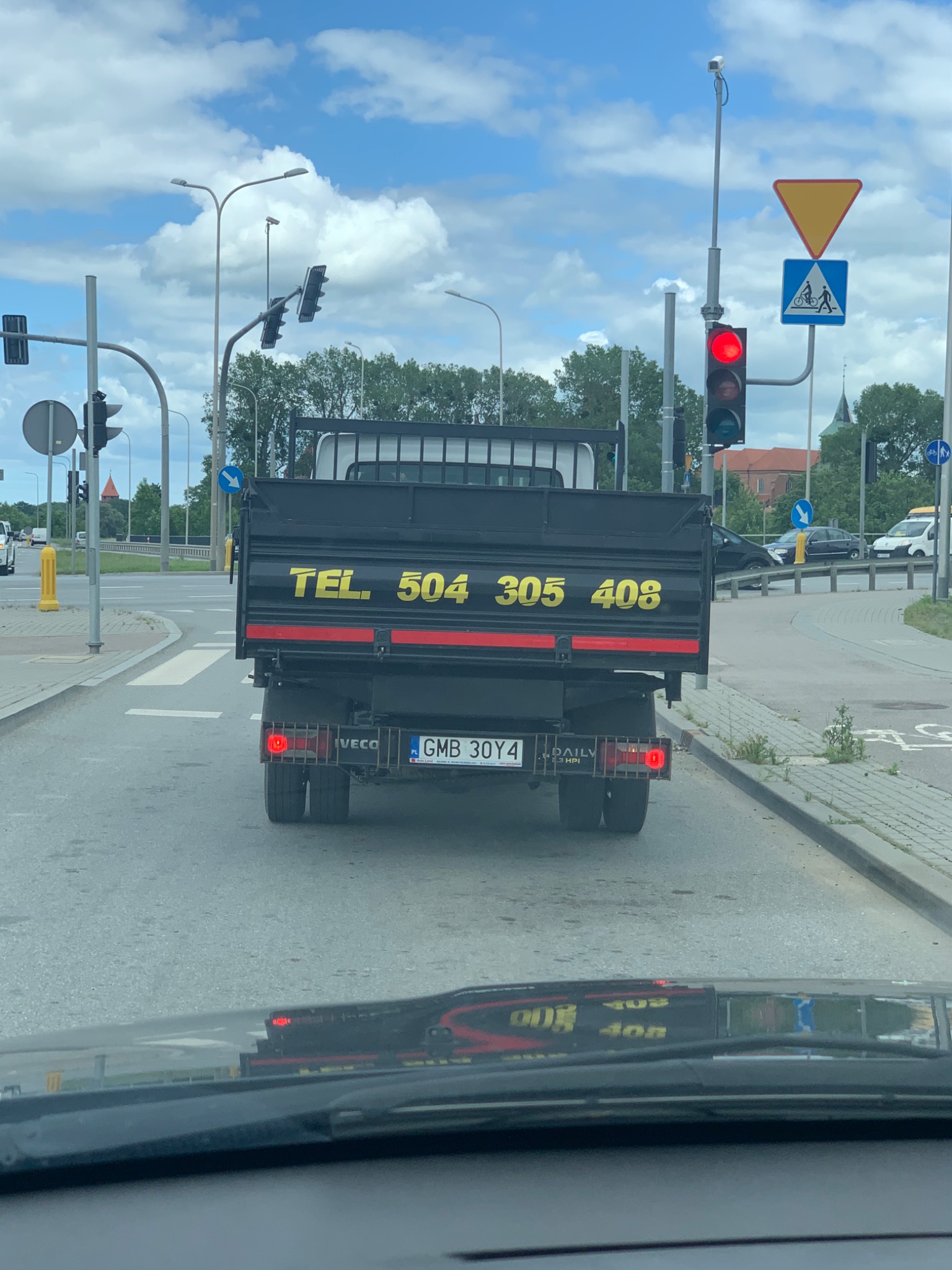 Ciężarówka Iveco z czarną skrzynią ładunkową i żółtym numerem telefonu na tylnej klapie, widziana z perspektywy kierowcy innego pojazdu na skrzyżowaniu z sygnalizacją świetlną.