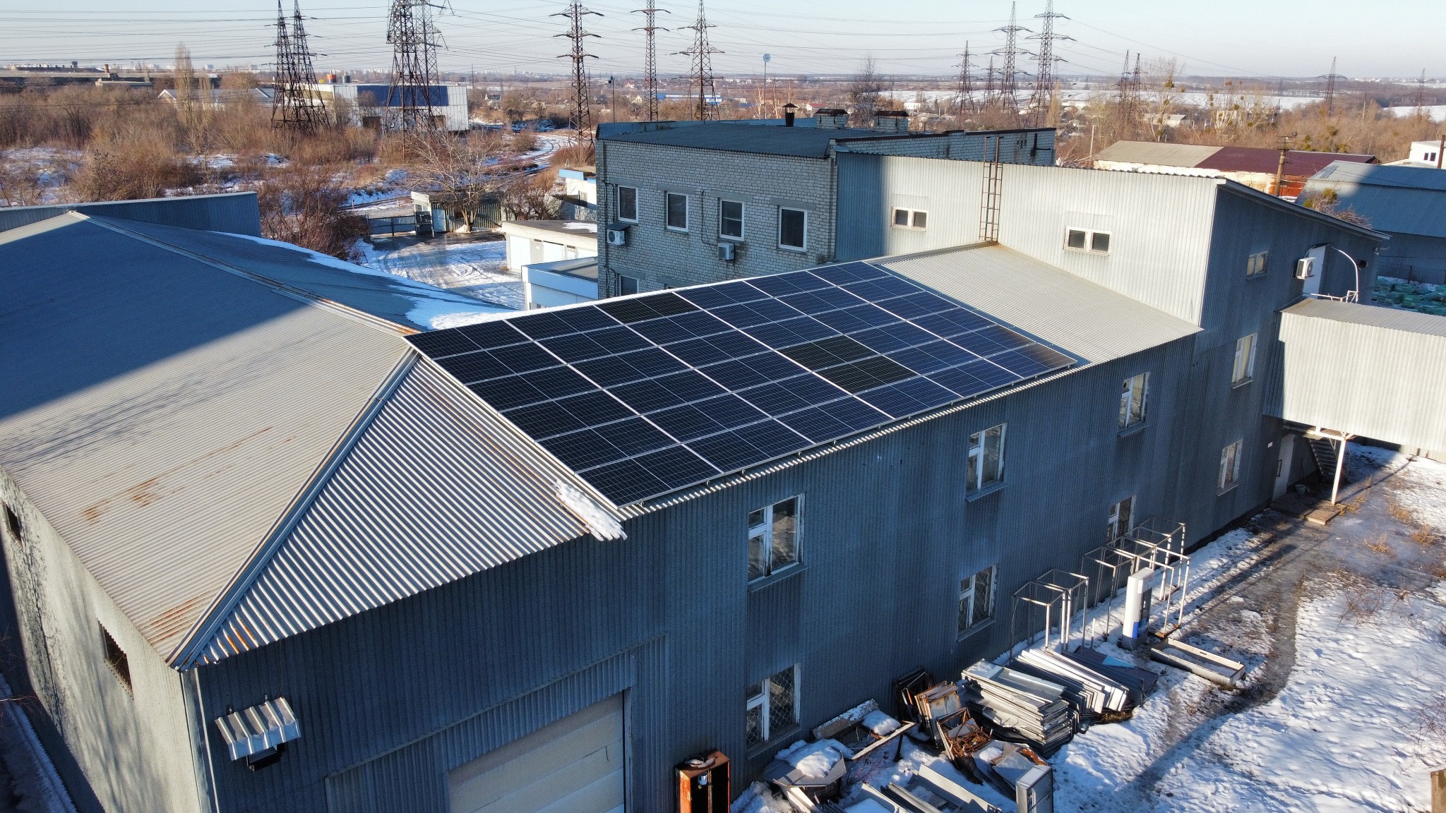 Instalacja paneli fotowoltaicznych na dachu budynku przemysłowego z blachy falistej, widok z góry na częściowo ośnieżony teren.