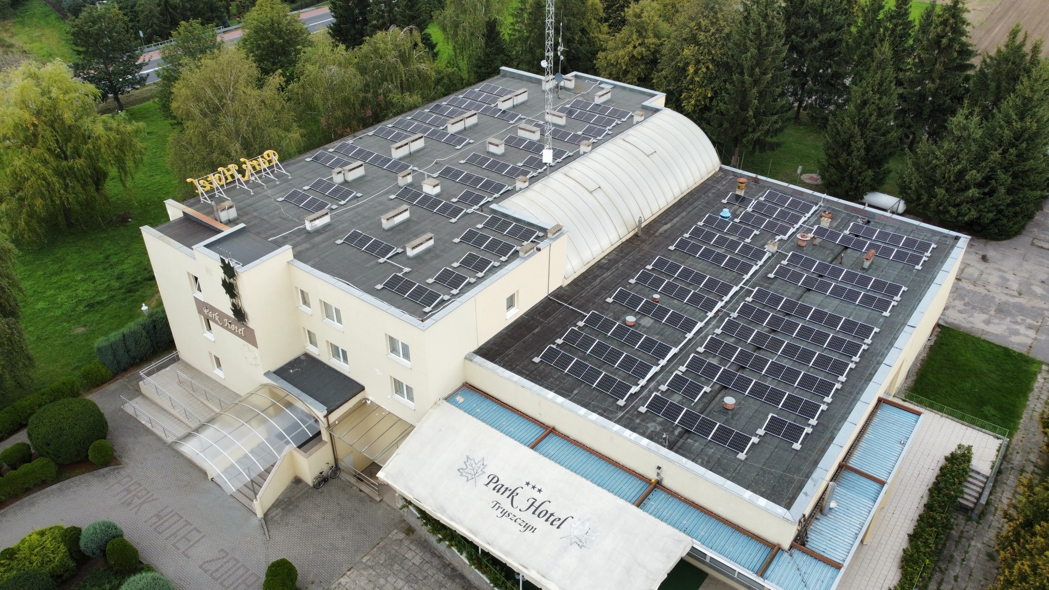 Widok z lotu ptaka na budynek hotelu z panelami słonecznymi na dachu, otoczony zielenią i drzewami.