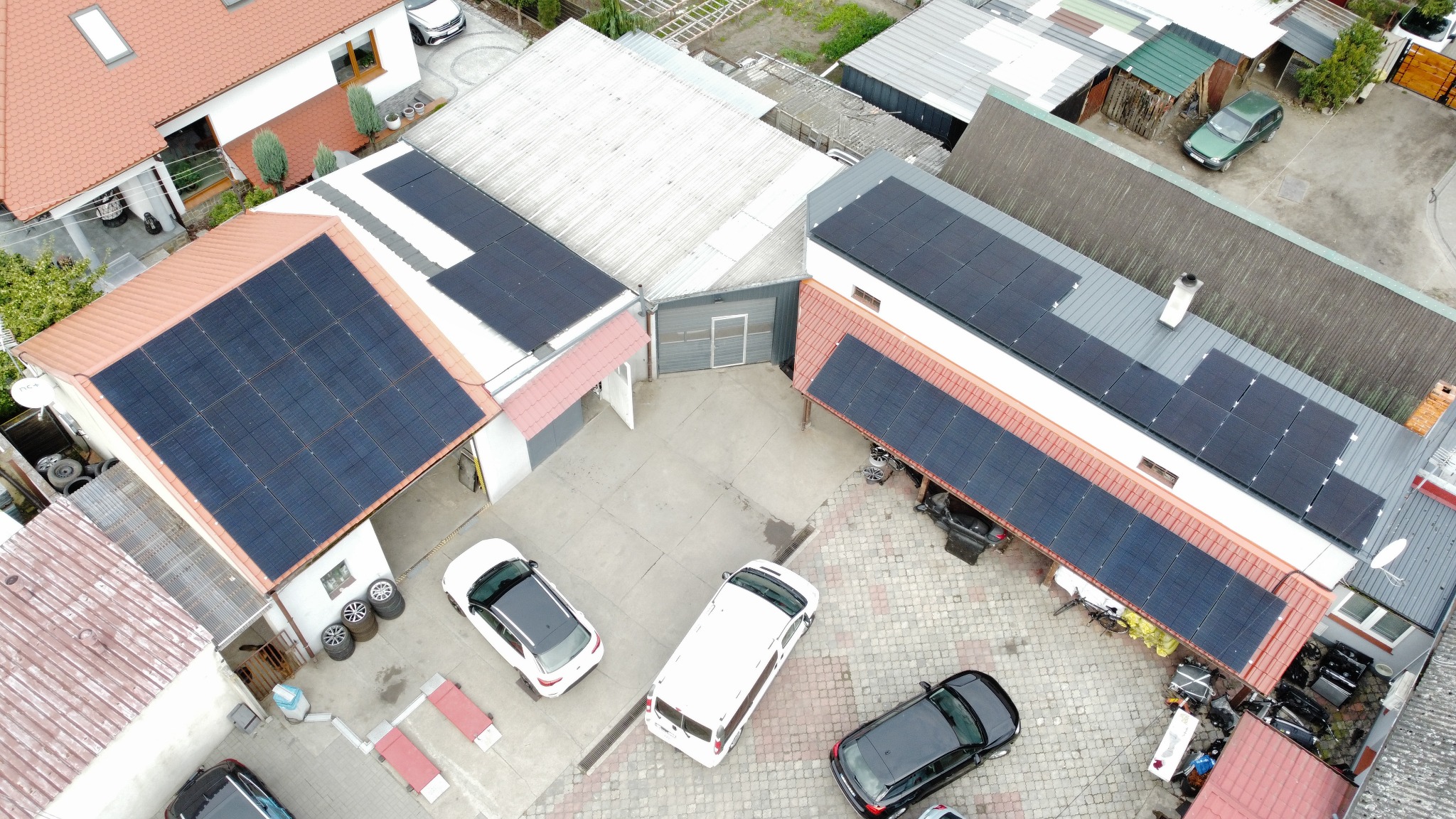 Widok z góry na budynki z panelami słonecznymi na dachach, zaparkowane samochody i podwórze z kostką brukową.