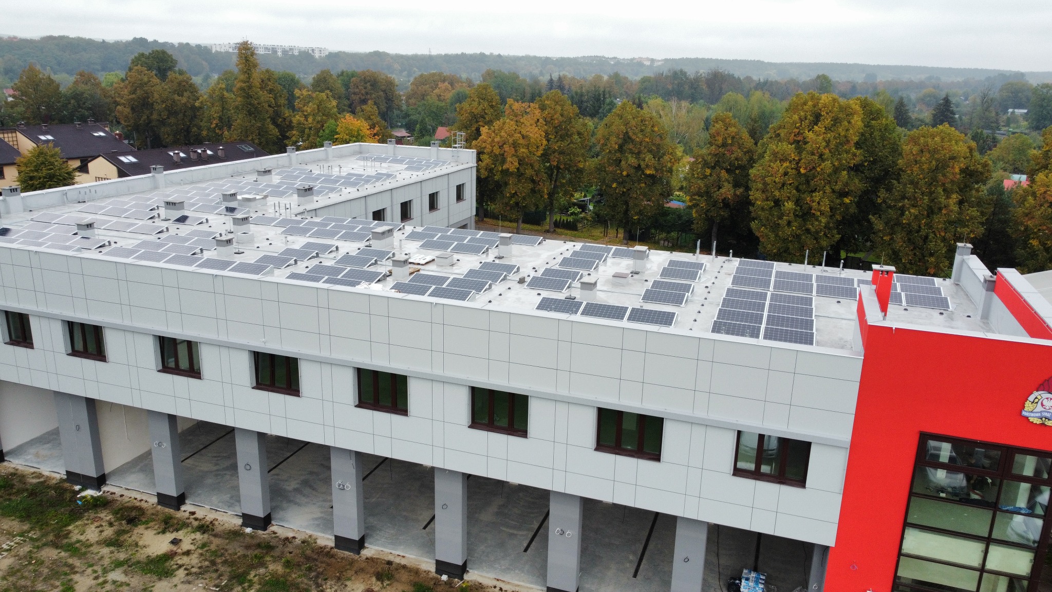 Widok z góry na budynek z zainstalowanymi panelami słonecznymi na dachu, otoczony drzewami w jesiennych barwach. Budynek posiada jasnoszarą elewację z ciemnymi oknami i czerwoną, wyróżniającą się...