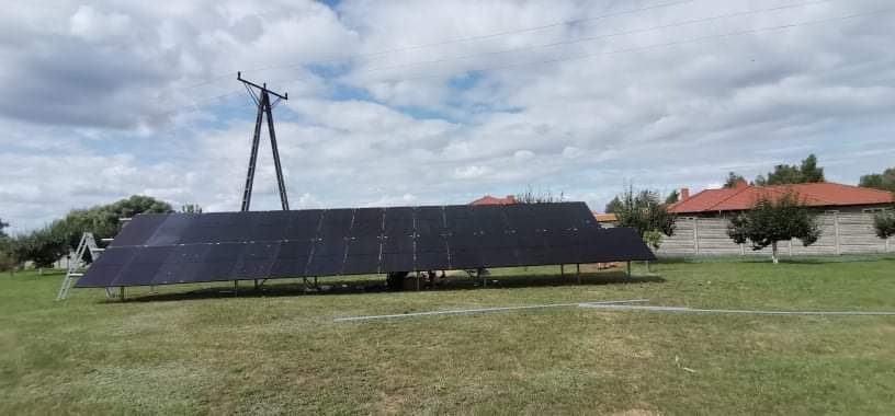 Nisko zawieszone panele słoneczne na trawiastym terenie, z widoczną drabiną i słupem elektrycznym w tle.