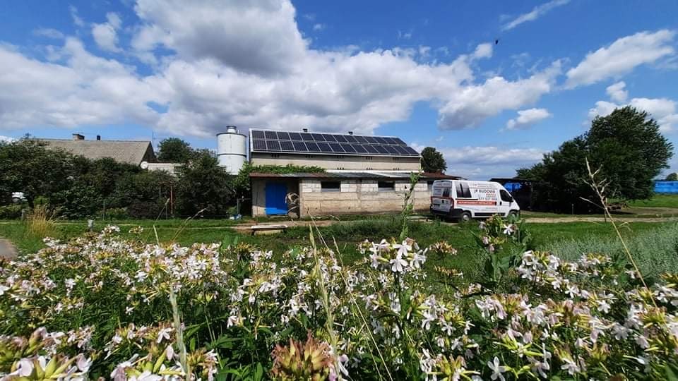 Panele słoneczne zamontowane na dachu budynku gospodarczego w wiejskim krajobrazie, z kwitnącymi kwiatami na pierwszym planie i samochodem serwisowym w tle.