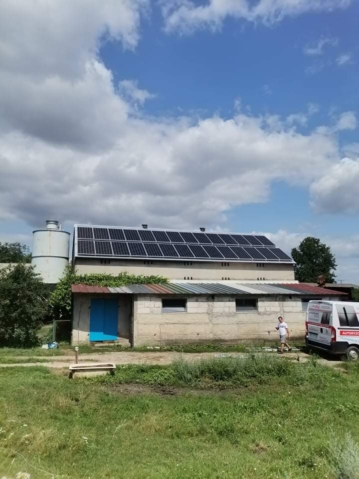 Panele słoneczne na dachu budynku gospodarczego z blaszanym dachem, obok silos, na pierwszym planie zarośnięty teren, osoba i samochód serwisowy w oddali.