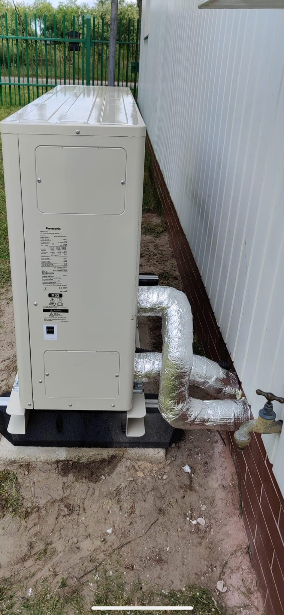 Zewnętrzna jednostka pompy ciepła Panasonic zainstalowana przy ścianie budynku z orurowaniem w izolacji aluminiowej i kranem na pierwszym planie.