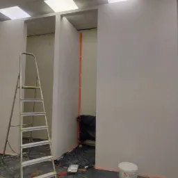 Wnętrze w trakcie odświeżania: świeżo pomalowane ściany na biało, zabezpieczone taśmą malarską, rozłożona folia ochronna, aluminiowa drabina oparta o ścianę, widoczne wiadro z farbą i narzędzia...