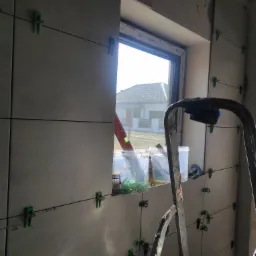 Świeżo ułożone, szare płytki ścienne z zielonymi klipsami poziomującymi, widoczne wiaderka z zaprawą i drabina, w tle okno z widokiem na dom.