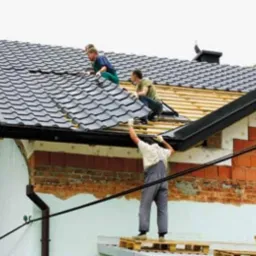 Trzech mężczyzn pracuje na dachu domu jednorodzinnego, układając szarą dachówkę. Jeden z nich stoi na paletach i podaje elementy, dwaj pozostali montują je na drewnianej konstrukcji. Widoczny...