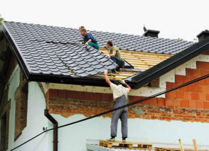 Trzech mężczyzn pracuje na dachu domu jednorodzinnego, układając szarą dachówkę. Jeden z nich stoi na paletach i podaje elementy, dwaj pozostali montują je na drewnianej konstrukcji. Widoczny...