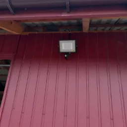 Zewnętrzny reflektor LED z czujnikiem ruchu zamontowany na bordowej ścianie z blachy trapezowej, widoczny brązowy system rynnowy.