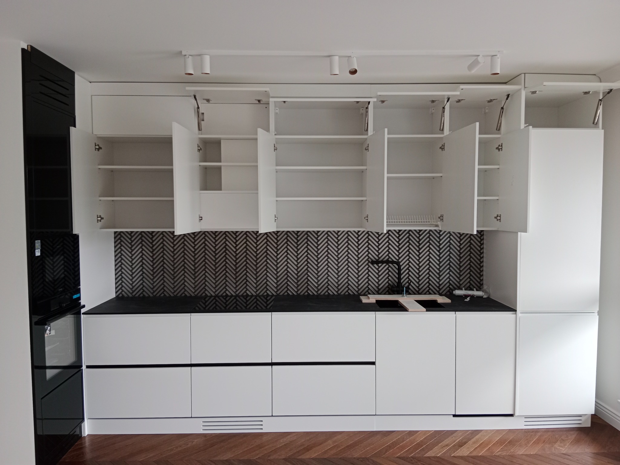 Nowoczesna biała zabudowa kuchenna z czarnym blatem i otwartymi szafkami górnymi, widoczna czarna armatura i płytki w jodełkę na ścianie, parkiet na podłodze.