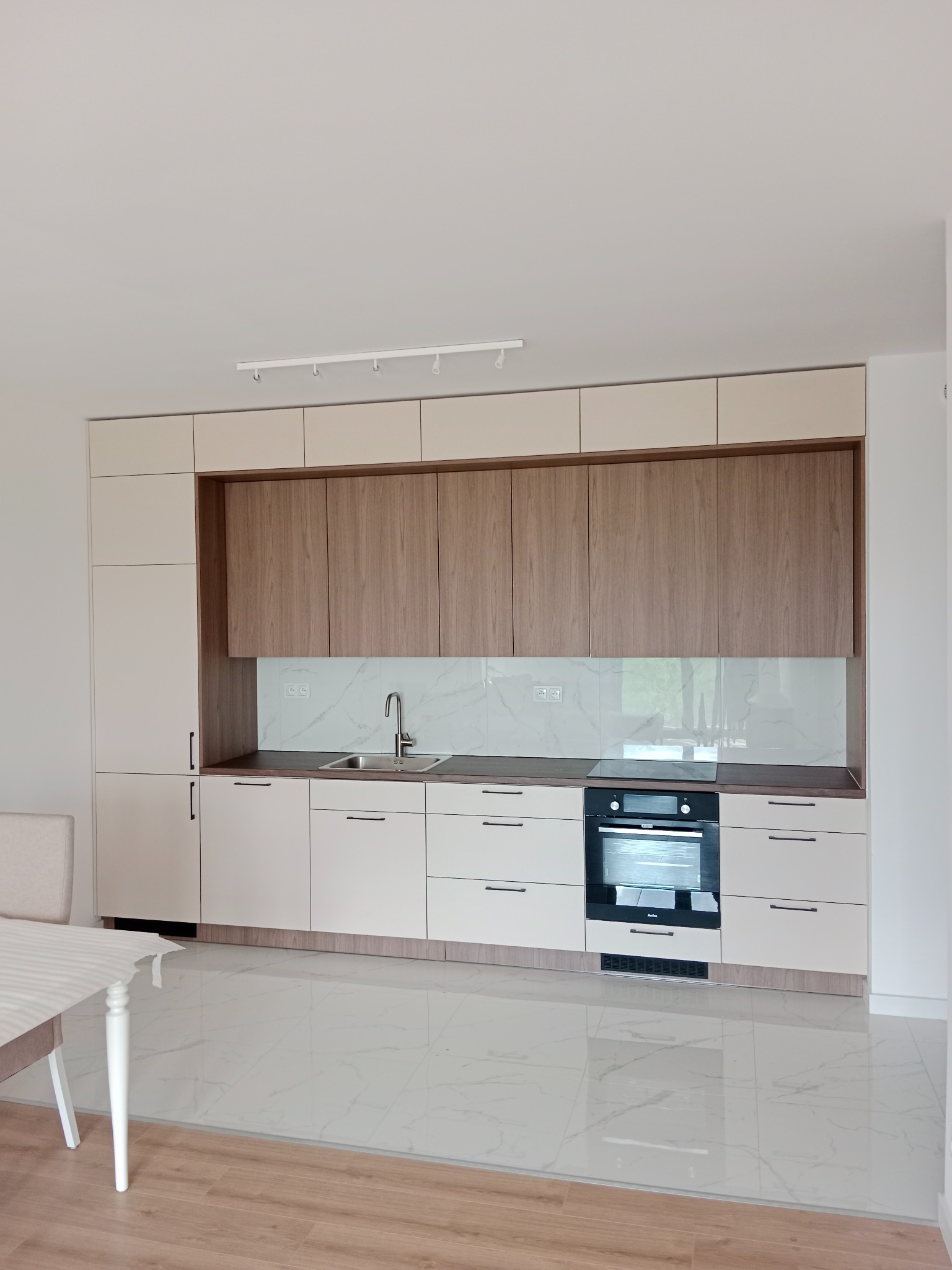 Nowoczesna kuchnia z beżowymi i drewnianymi szafkami, marmurowy backsplash, płyta indukcyjna i piekarnik w zabudowie, biała podłoga z marmurowym wzorem, stół z krzesłem w rogu.