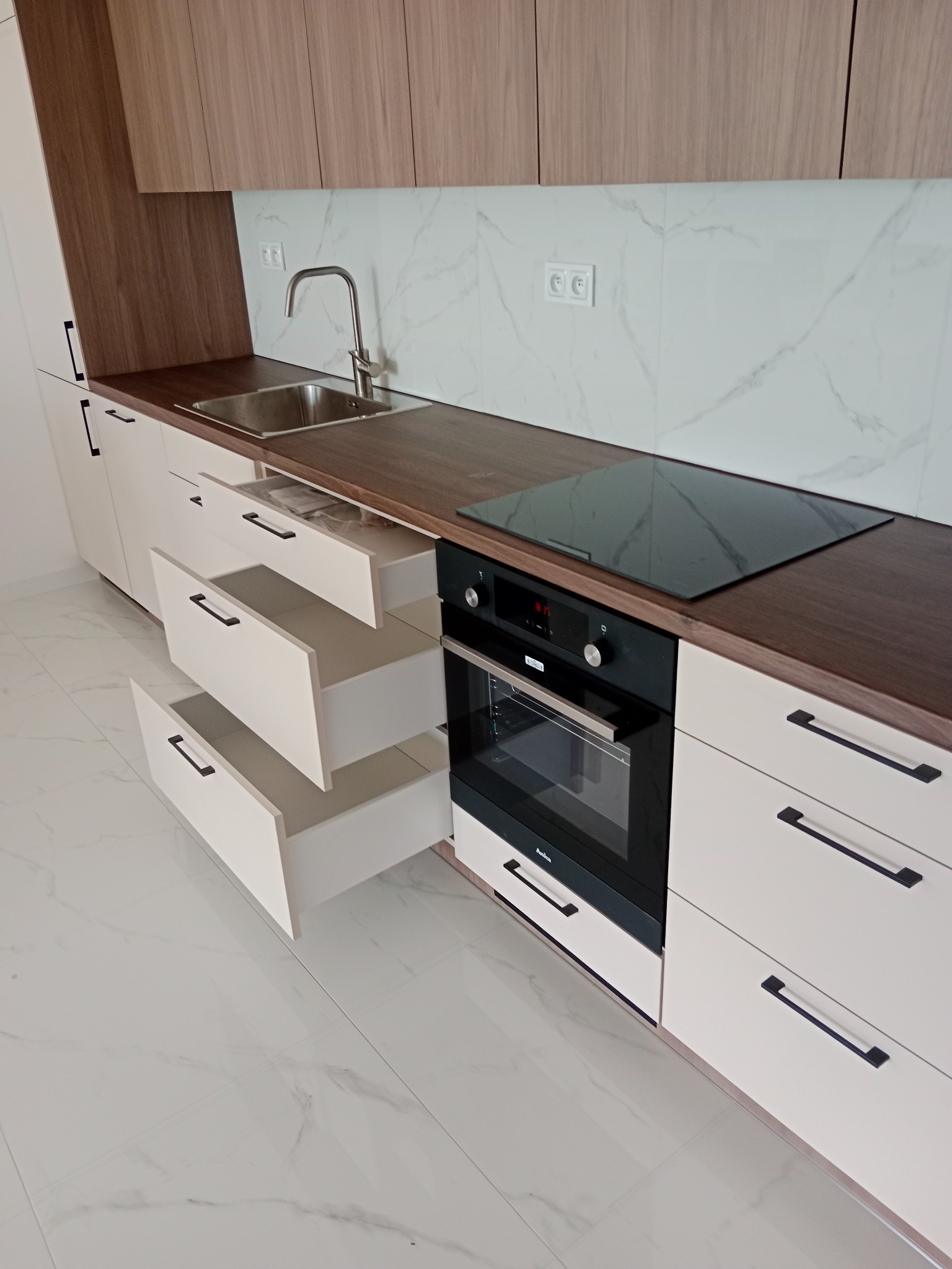 Nowoczesna kuchnia z otwartymi szufladami, brązowym blatem i płytą indukcyjną, białe fronty szafek z czarnymi uchwytami, marmurowe płytki na ścianie.