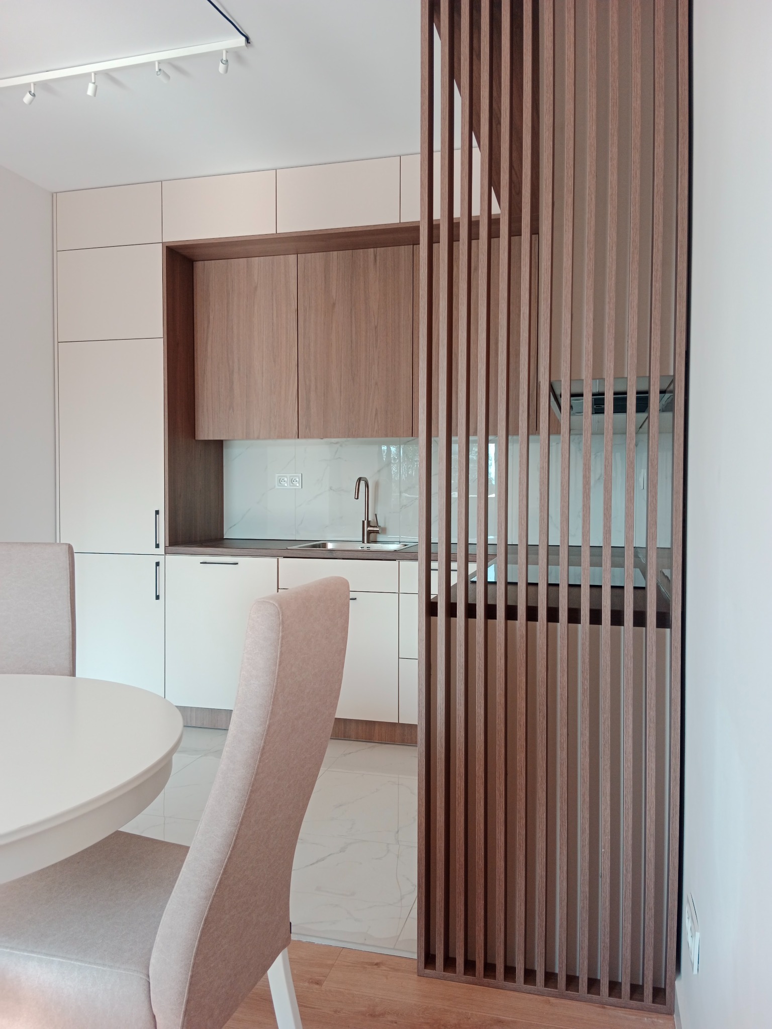 Nowoczesna kuchnia z białymi i drewnianymi szafkami, oddzielona od jadalni dekoracyjną, pionową, drewnianą ścianką działową, widoczne dwa krzesła przy okrągłym stole.