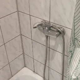 Biała wanna z chromowaną baterią i słuchawką prysznicową na tle białych płytek z szarym wzorem marmuru, częściowo zasłonięta zasłoną w zielone liście. Widoczne ślady osadu na fugach.