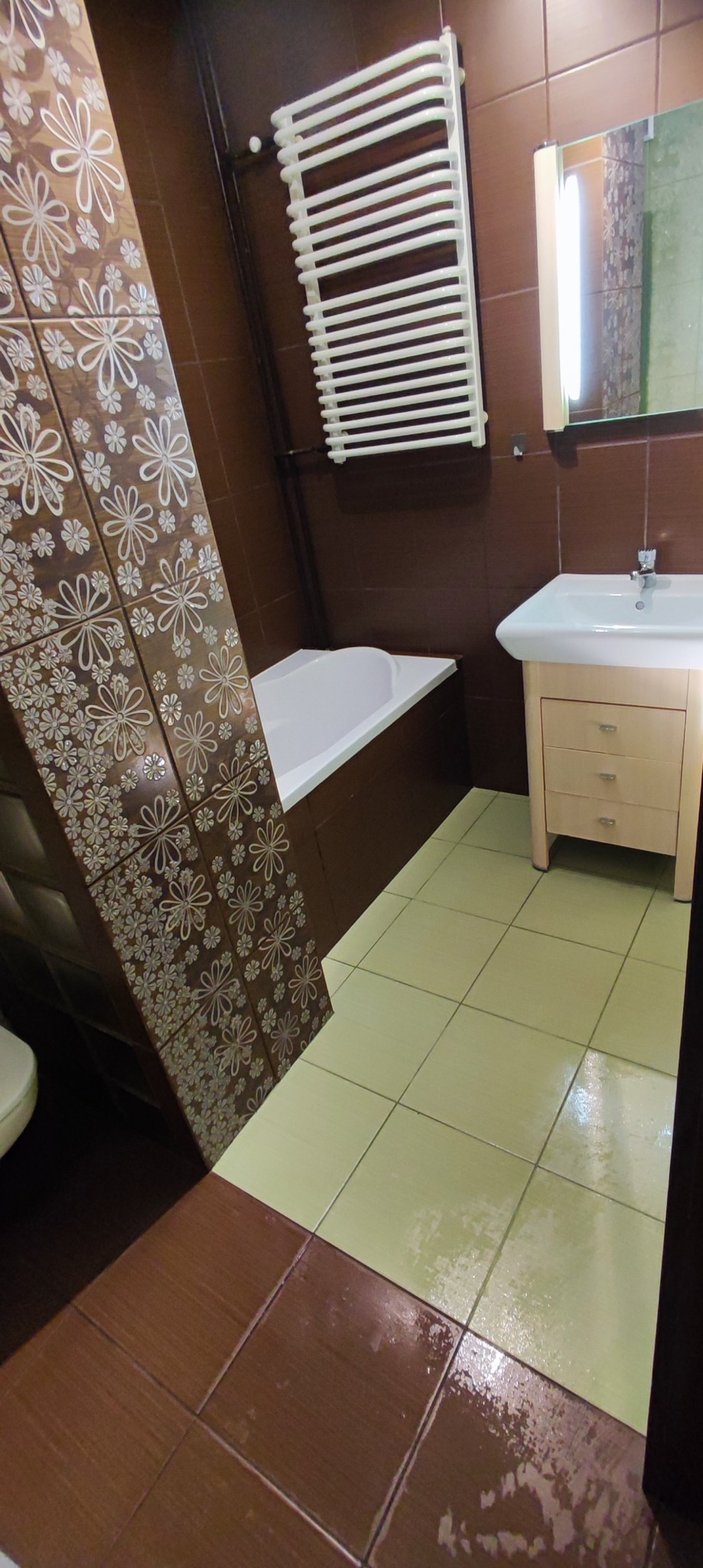 Ukośne ujęcie wykafelkowanej łazienki z brązowymi i beżowymi płytkami, białą wanną, grzejnikiem drabinkowym i umywalką z szafką, podłoga częściowo mokra.