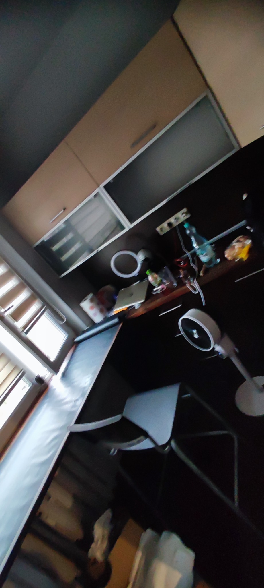 Niezbyt ostre ujęcie pod kątem na zagracony blat kuchenny z butelką wody, laptopem, wentylatorem i innymi przedmiotami, widoczne szafki kuchenne i fragment okna z żaluzjami.