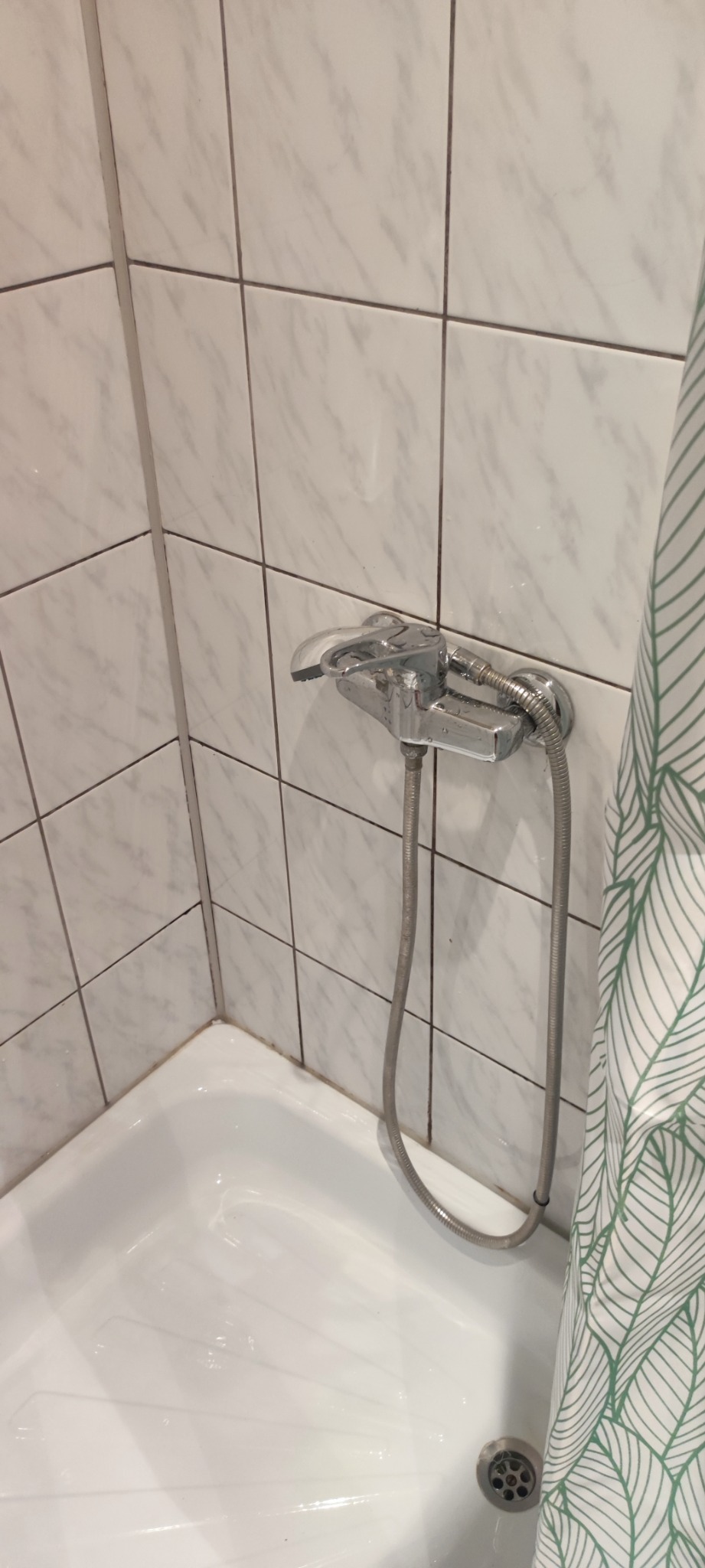 Biała wanna z chromowaną baterią i słuchawką prysznicową na tle białych płytek z szarym wzorem marmuru, częściowo zasłonięta zasłoną w zielone liście. Widoczne ślady osadu na fugach.