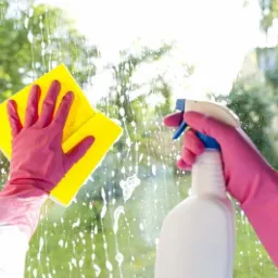 Mycie okna: ręce w różowych rękawicach spryskują szybę płynem i wycierają żółtą ściereczką na tle zieleni drzew.