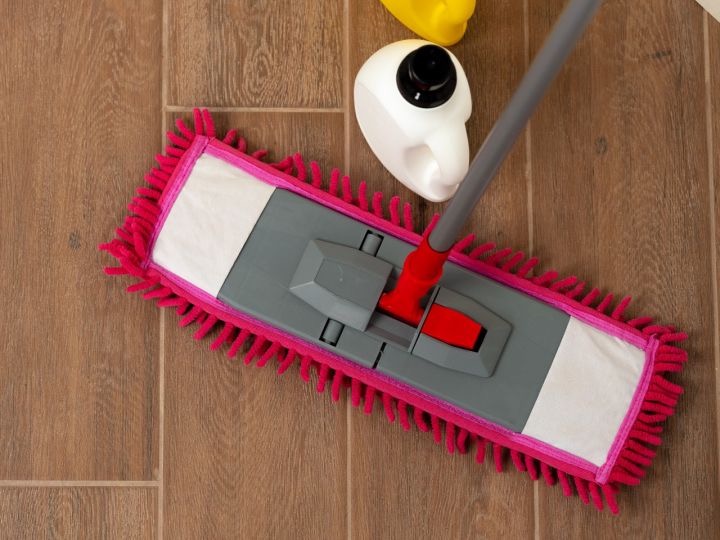 Mop płaski z różową obwódką i szarym korpusem na panelach podłogowych, obok płyn do czyszczenia w białej butelce i żółty pojemnik.
