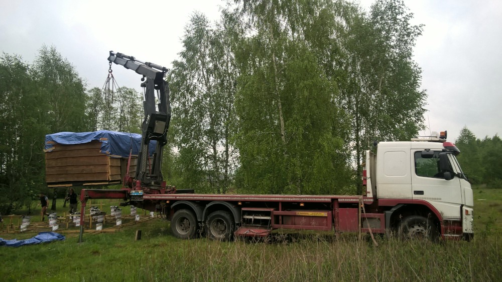 Ciężarówka z dźwigiem przenosi drewnianą konstrukcję, prawdopodobnie moduł domu, na przygotowany fundament w lesistym terenie.