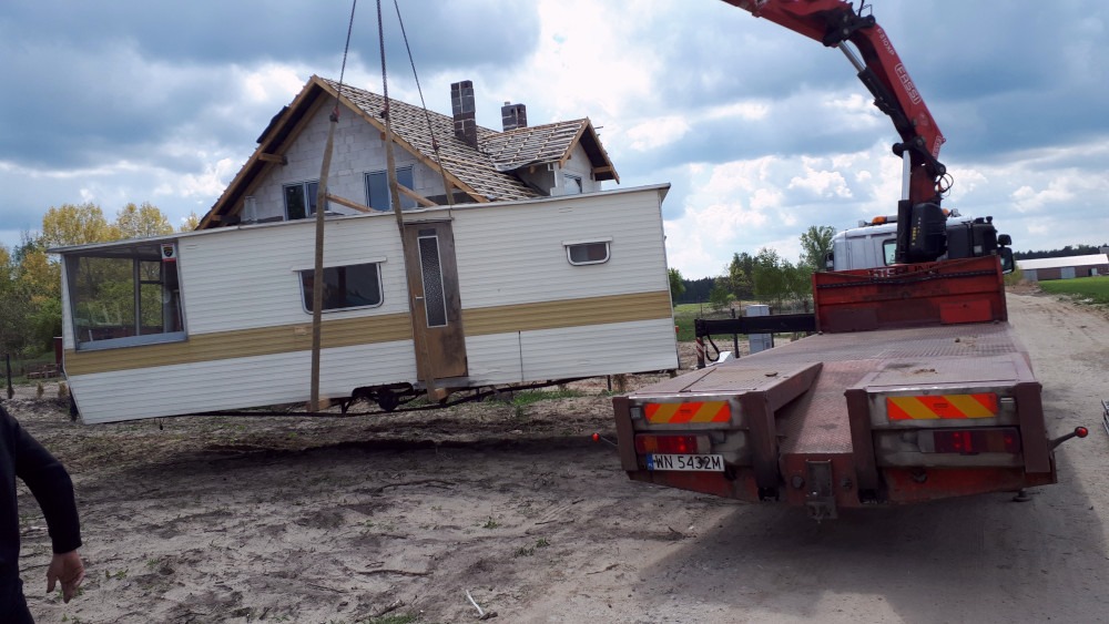 Transport mobilnego domu z werandą na platformie ciężarówki z dźwigiem na tle budowy domu jednorodzinnego.