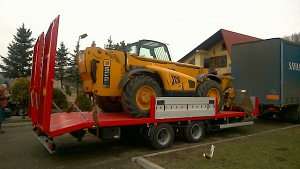 Żółta ładowarka teleskopowa JCB 532 zabezpieczona łańcuchami na czerwonej niskopodwoziowej przyczepie transportowej, z tyłu zaparkowana naczepa ciężarówki.