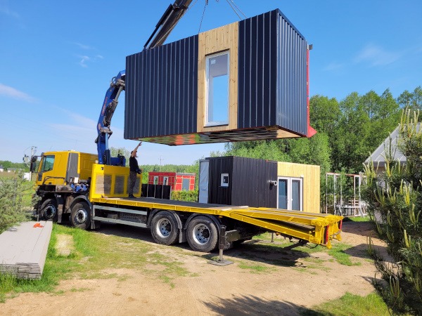 Żółty samochód ciężarowy z dźwigiem podnosi modułowy dom z oknem, w tle inne domki modułowe i drzewa.