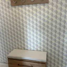 Drewniany panel z czterema haczykami i półką powyżej, oraz skrzynia z siedziskiem w przedpokoju na tle ściany w geometryczne wzory.