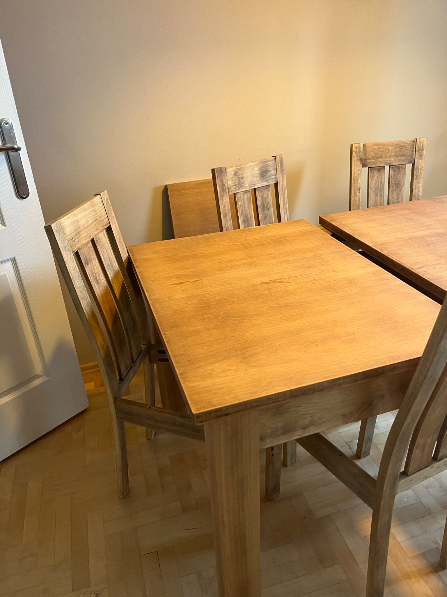 Rozkładany stół z krzesłami z jasnego drewna, widoczny fragment podłogi z parkietu i białe drzwi z chromowaną klamką.