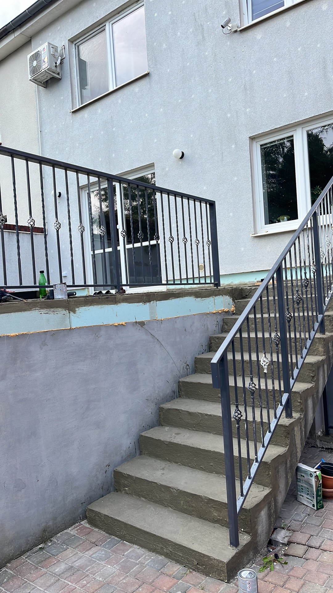 Schody zewnętrzne z metalową balustradą o ozdobnych elementach, prowadzące do drzwi wejściowych budynku z widoczną izolacją termiczną fundamentów.