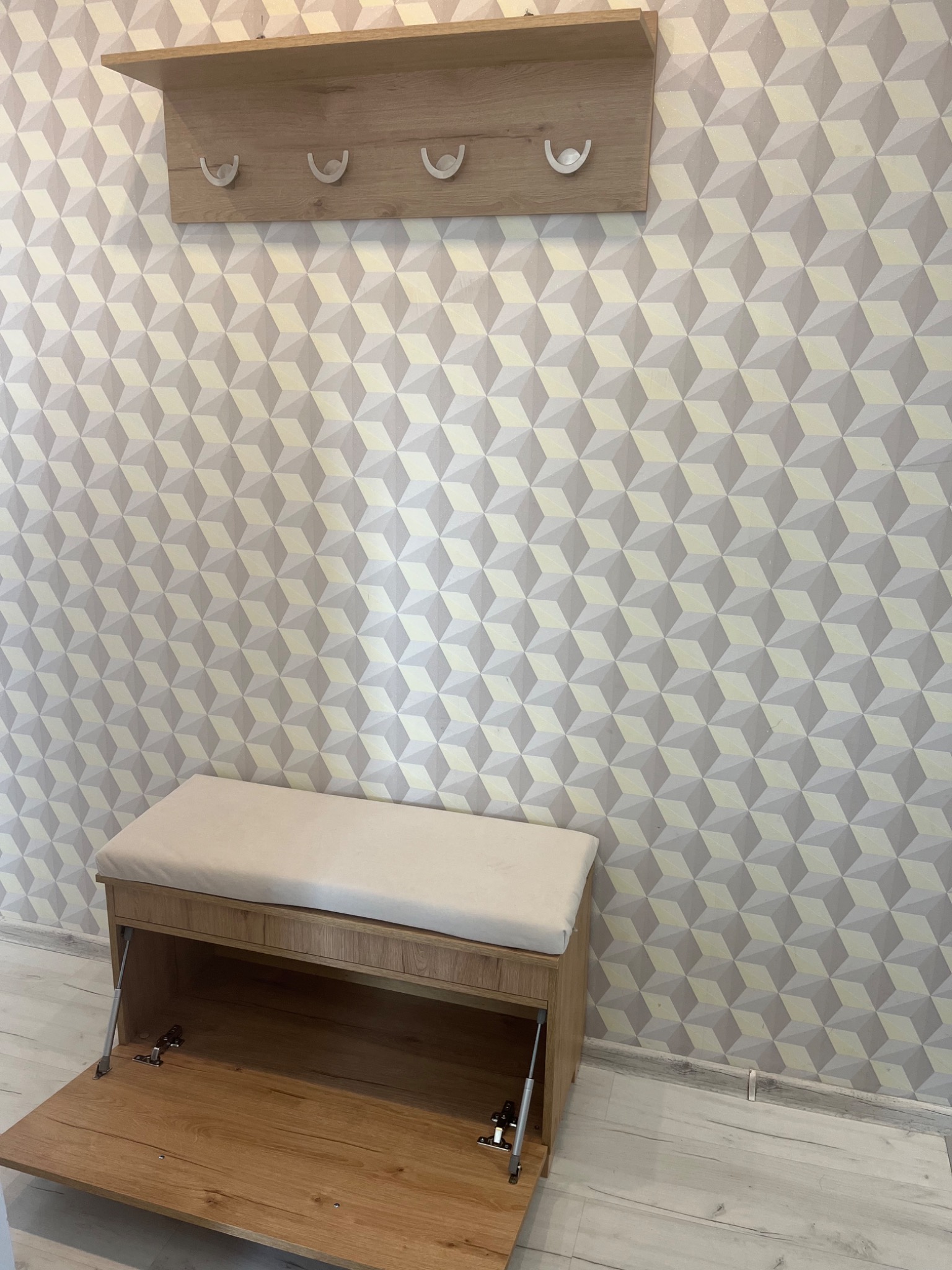 Drewniana szafka z siedziskiem i otwartym schowkiem, zawiasy widoczne, nad nią wieszak z półką, tapeta w geometryczne wzory w tle.