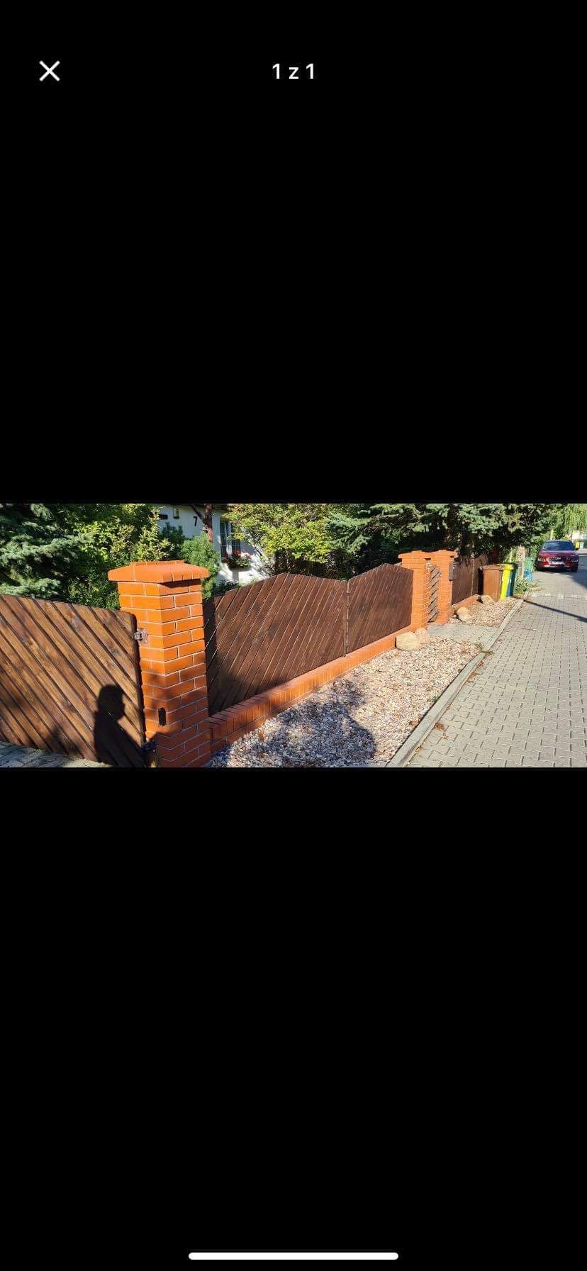 Nowoczesne ogrodzenie z brązowych desek ułożonych ukośnie, wsparte na ceglanych słupkach, oddzielające posesję od chodnika wyłożonego kostką.