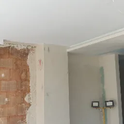 Wnętrze w trakcie remontu, widoczna ściana z cegły częściowo pokryta tynkiem, gładź na ścianach i suficie, lampa budowlana na statywie.