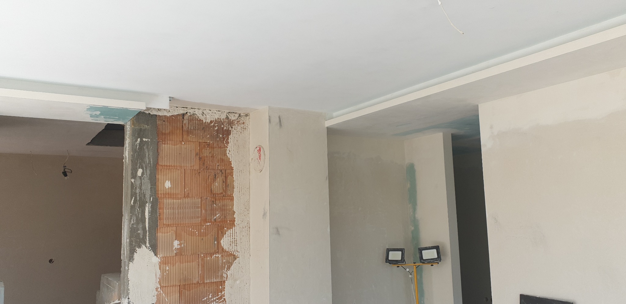 Wnętrze w trakcie remontu, widoczna ściana z cegły częściowo pokryta tynkiem, gładź na ścianach i suficie, lampa budowlana na statywie.