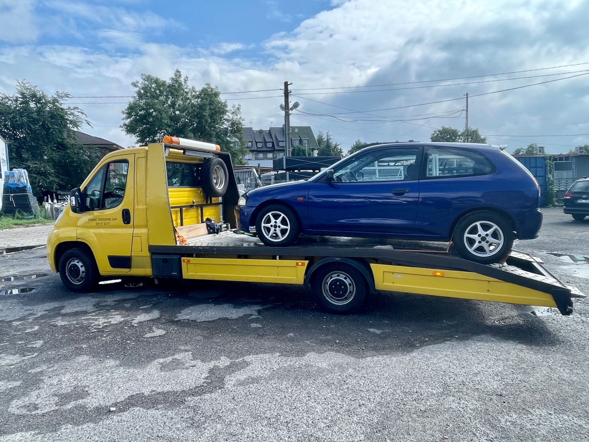 Żółta laweta z niebieskim samochodem osobowym na platformie, zaparkowana na asfaltowym placu z budynkami w tle i pochmurnym niebem.
