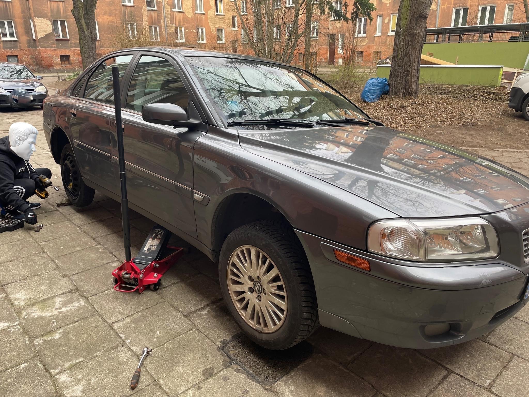 Szary samochód osobowy na podnośniku hydraulicznym, obok osoba w masce wymienia koło, tło stanowi budynek mieszkalny.