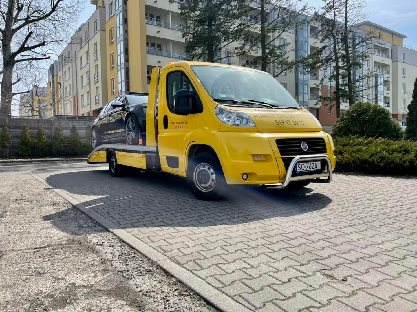 Żółta laweta Fiat z czarnym samochodem osobowym na platformie, zaparkowana na chodniku z kostki brukowej przed budynkiem mieszkalnym w słoneczny dzień. Widoczny numer telefonu alarmowego na kabinie...