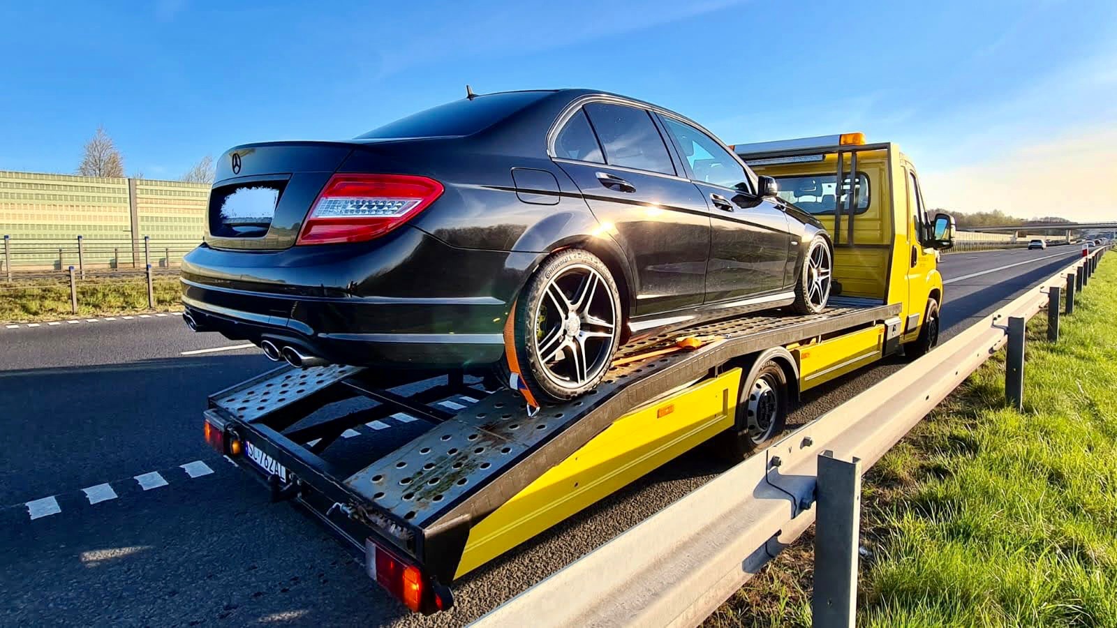 Czarny samochód osobowy marki Mercedes zabezpieczony pasami na żółtej lawecie, widok z tyłu na autostradzie w słoneczny dzień.