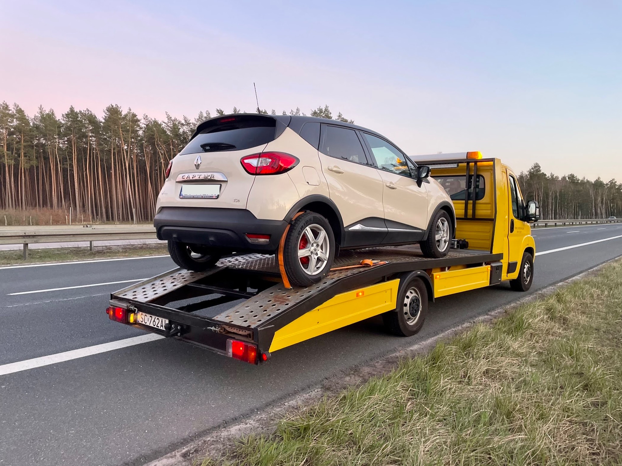 Beżowy Renault Captur przewożony na żółtej lawecie wzdłuż drogi, widok z tyłu, las w tle.