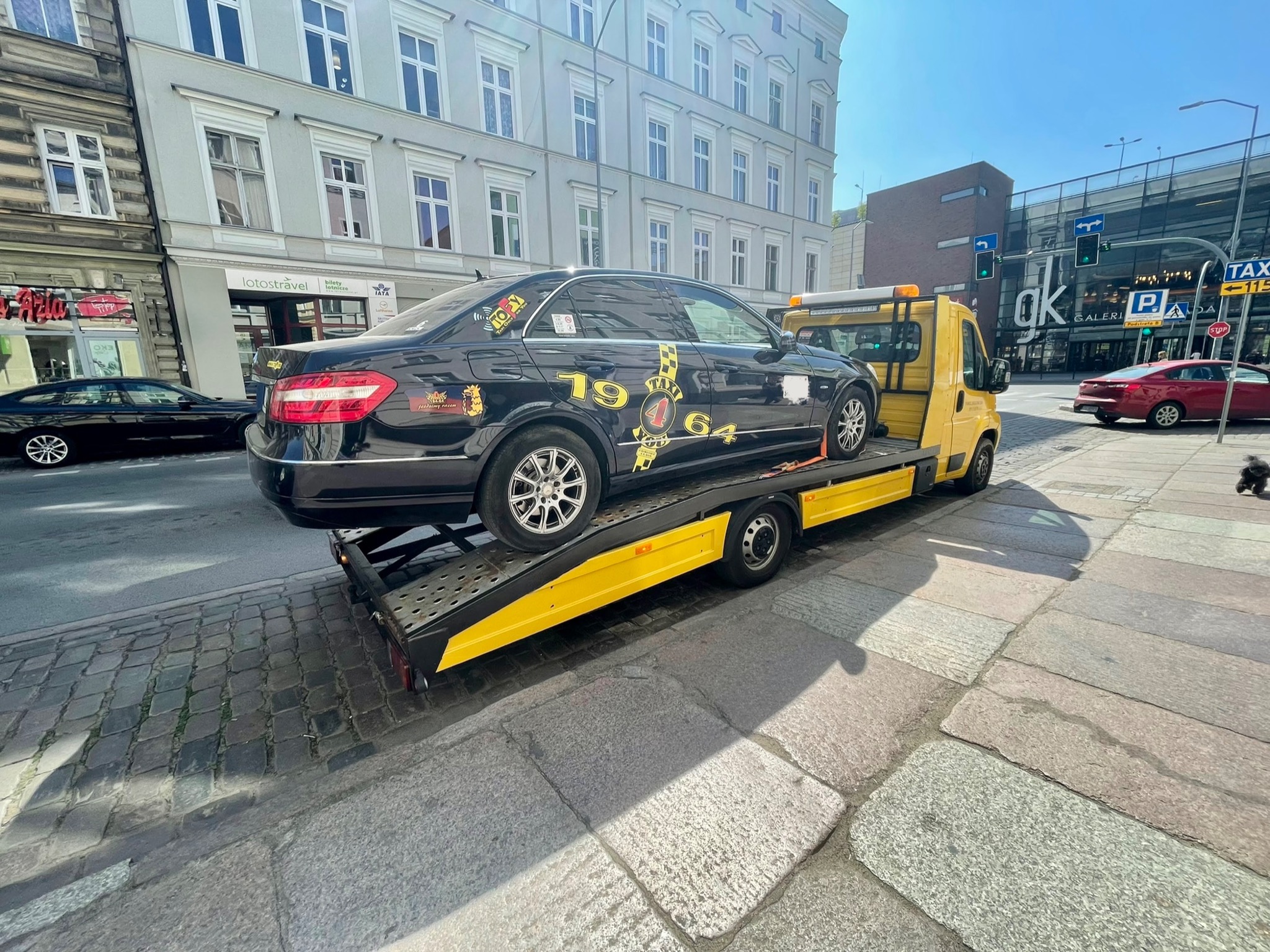 Czarny samochód osobowy z naklejkami na karoserii przewożony na żółtej lawecie na tle miejskiej ulicy z budynkami i sygnalizacją świetlną.