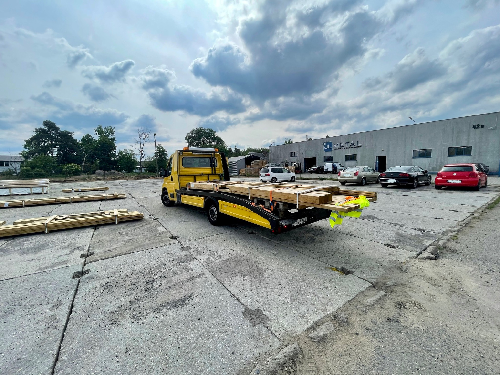Żółta laweta z drewnem na platformie, zaparkowana na betonowym placu przed budynkiem firmy JL Metal, pochmurne niebo w tle.